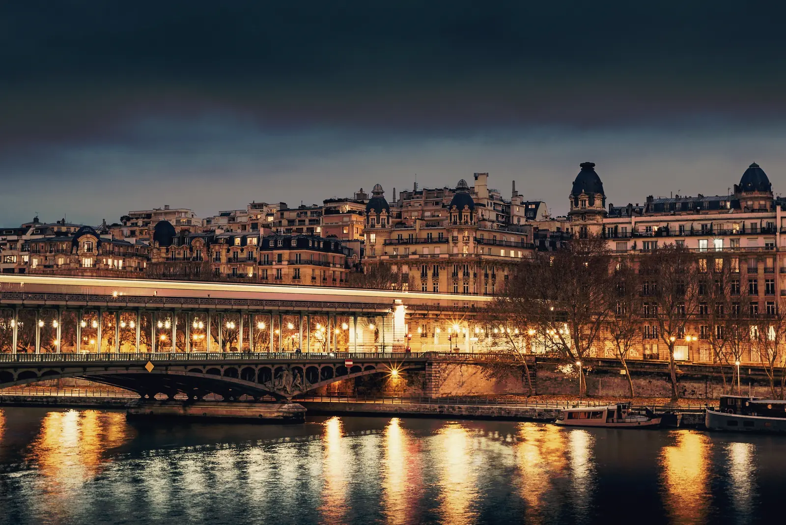 A stone bridge and river, old apartment buildings lit at dusk.
