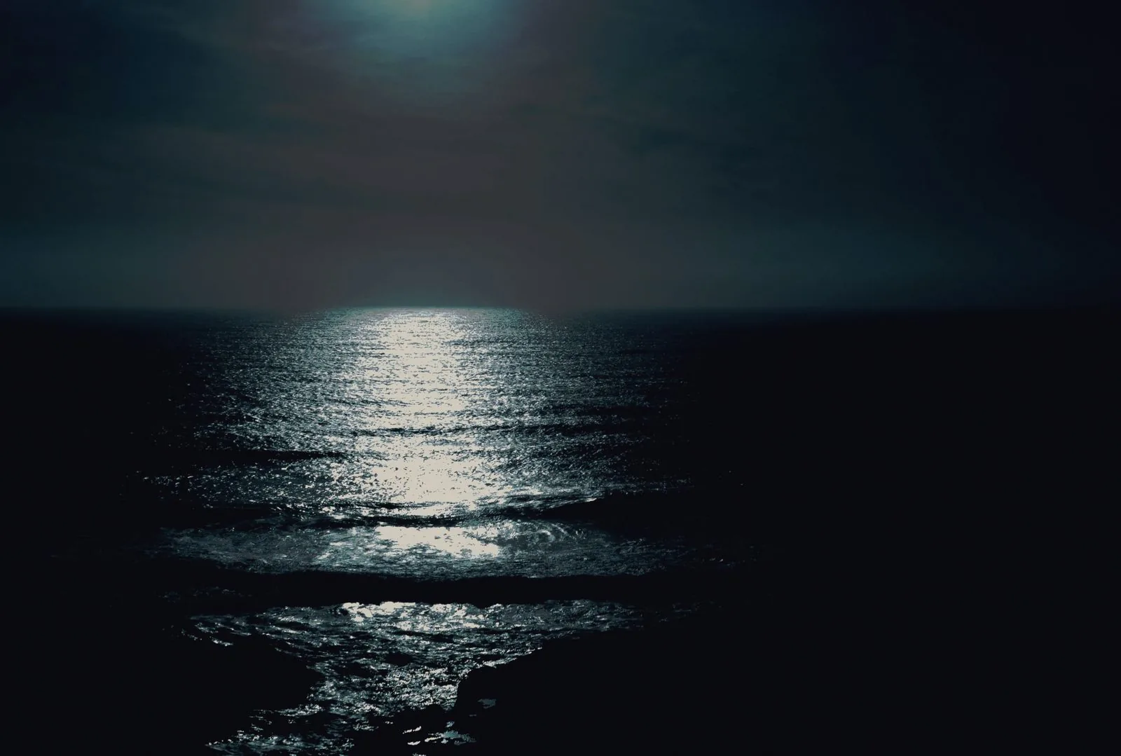 Open water at night under heavy cloud, one path of light across it.