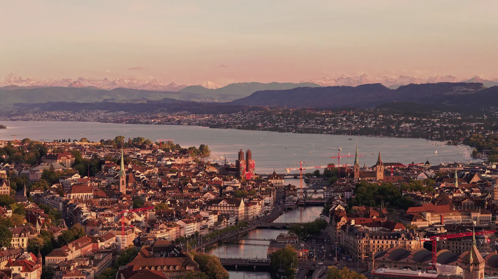 Zurich&rsquo;s old town seen from height across the lake at dusk, the Alps on the horizon.